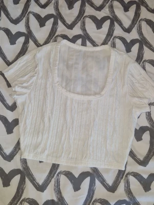 Croptop - Snygg vit croppad topp med korta ärmar och fyrkantig ringning. Tillverkad i ett strukturerat, lätt material som ger en avslappnad och trendig look. Perfekt att matcha med höga jeans eller kjol.