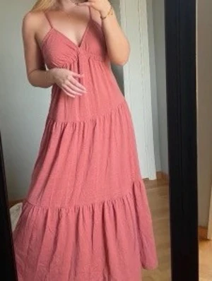 Lång rosa sommarklänning med smala axelband - Luftig långklänning i rosa med smala axelband och v-ringad hals. Klänningen har volangdetaljer och är tillverkad i ett lätt material, perfekt för varma dagar. Passformen är loose och modellen är ärmlös.