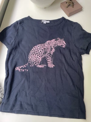 Mörkblå t-shirt med rosa leopard - Cool mörkblå t-shirt med ett stort rosa leopardtryck på framsidan. Klassisk rund halsringning och korta ärmar. Tillverkad i mjuk bomull som känns skön mot huden. Perfekt för dig som gillar djurmotiv och vill sticka ut lite extra. Använt en gång!