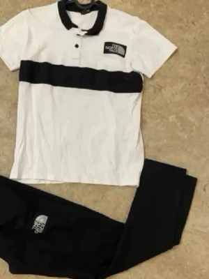 The North Face svartvitt set - Snyggt set från The North Face med en vit pikétröja med svart krage, knappslå och bred svart rand över bröstet samt matchande svarta byxor. Båda plaggen har tydliga logotyper. Perfekt för en sportig och avslappnad look.