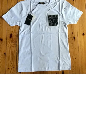 Vit Fendi t-shirt med logoficka - Vit t-shirt från Fendi med bra passform. På bröstet finns en svart logoficka med Fendis  mönster. Perfekt för dig som vill ha en lyxig look. Materialet är mjuk bomull. Om ni vill ha bättre eller fler bilder säg till 