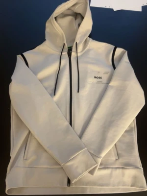 Vit hoodie med dragkedja från BOSS - Snygg vit hoodie från BOSS med dragkedja framtill och svarta detaljer vid axlarna. Huva med svarta snören och två fickor på sidorna. Tillverkad i mjukt material som känns skönt mot huden. Liten BOSS-logga på bröstet för en clean look.