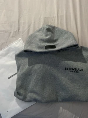 Grå Essentials Fear of God hoodie - Snygg grå hoodie från Essentials Fear of God med svart logga på bröstet och patch på huvan. Klassisk passform med känguruficka och ribbade muddar. Perfekt för en avslappnad streetwear-look. Tillverkad i mjukt bomullsmaterial.