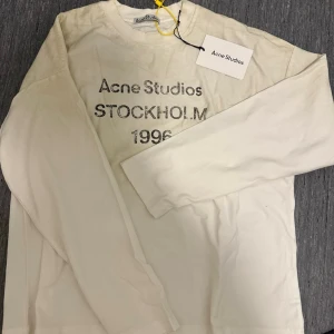 Vit långärmad topp Acne Studios Stockholm  - Vit långärmad topp från Acne Studios med tryck 'Acne Studios STOCKHOLM 1996' på bröstet. Klassisk rund halsringning och mjukt bomullsmaterial. Perfekt för dig som gillar stilrena och trendiga plagg med ikonisk branding.