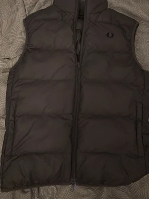 Svart dunväst från Fred Perry - Snygg svart dunväst från Fred Perry med diskret logga på bröstet. Västen har hög krage, dragkedja framtill och quiltad design som ger en puffig look. Perfekt att slänga över en hoodie eller t-shirt för extra värme. Materialet är slitstarkt och håller värmen bra.