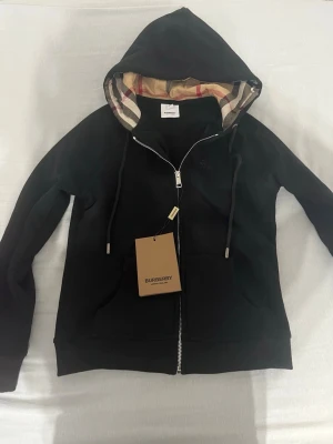 Svart hoodie från Burberry med dragkedja - Helt ny Stilren svart hoodie från Burberry med dragkedja och klassiskt rutigt foder i huvan. Hoodien har dragsko vid huvan och ribbade muddar. Märkeslapp kvar på plagget. Liten i storleken 