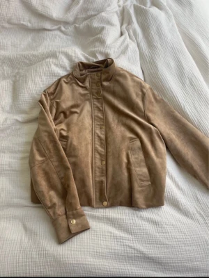 Beige bomberjacka i mockaimitation - Snygg beige bomberjacka i mockaimitation från Zara med ståkrage och guldfärgade tryckknappar. En knapp har tappats, (bild 2) men annars superfint skick!! supersnygg men tyvärr lite kort i ärmarna på mig☺️