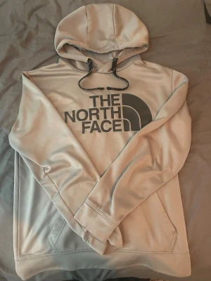 Grå hoodie från The North Face med luftträdande material - Snygg grå hoodie från The North Face med stor logga på bröstet. Hoodien har huva med svarta dragsnören och en klassisk känguruficka framtill. Tillverkad i mjukt material som känns skönt mot huden. Perfekt för en avslappnad och trendig stil.