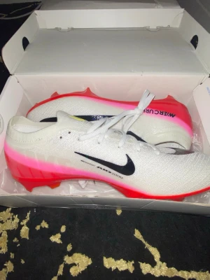 Nike Mercurial fotbollsskor vit/röd - Säljer ett par Nike Mercurial fotbollsskor i vitt med svart swoosh och röda detaljer. Skorna har en modern, strömlinjeformad design och är tillverkade i ett lätt syntetmaterial för bästa känsla på planen. Perfekta för dig som vill ha snabbhet och kontroll.