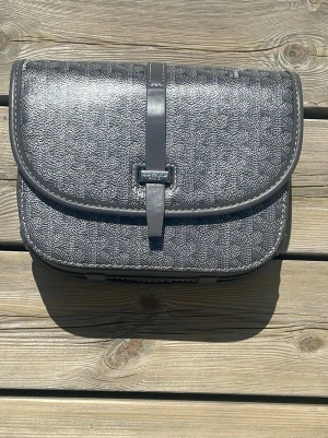 Grå väska med mönster - Snygg grå handväska  med klassiskt vitt mönster och rundade kanter. Väskan har ett lock med rem och spänne framtill samt en praktisk ficka på baksidan. Tillverkad i canvas med läderdetaljer och tydlig logotyp på baksidan.