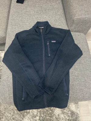 Blå fleecejacka från Patagonia - Mysig blå fleecejacka från Patagonia med hel dragkedja framtill, hög krage och tre fickor med dragkedja. Perfekt för lager-på-lager och har en stilren look med diskret logga på bröstet. Passar både till chill och äventyr.