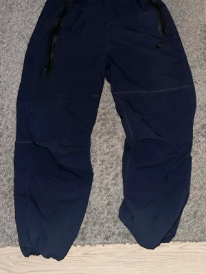 Mörkblå mjukisbyxor från Nike - Säljer ett par mörkblå joggers ifrån Nike, med resår i benslut och midja. Byxorna har svarta dragkedjor på fickorna och en avslappnad passform. Perfekt att ha på gymmet