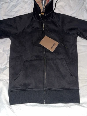 Svart hoodie från Burberry med dragkedja - Svart hoodie från Burberry med dragkedja framtill, klassisk rutmönstrad insida i huvan och stora fickor på magen. Justerbar huva med snören och ribbade muddar. Perfekt för dig som vill ha en stilren och lyxig look. Den är liten i size typ som S 