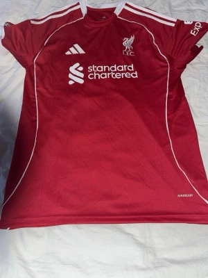 Liverpool matchtröja Szoboszlai #8 - Röd Liverpool FC matchtröja med vita detaljer, Adidas-logga och Standard Chartered-tryck på bröstet. Szoboszlai och nummer 8 på ryggen. Tillverkad i lätt och ventilerande material med Aeroready-teknologi. Perfekt för fotbollsträning eller att bära på matchdag.