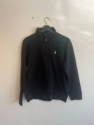 Svart half-zip hoodie från Polo Ralph Lauren - Svart hoodie från Polo Ralph Lauren med half-zip framtill och klassisk vit logga broderad på bröstet. Tillverkad i mjukt bomullsmaterial och har en hög krage. Perfekt för en clean och stilren look. (obs ej orginal)