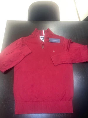 Röd stickad tröja Tommy Hilfiger - Snygg röd stickad tröja från Tommy Hilfiger med halv dragkedja och ribbade muddar. Tröjan har en liten broderad logga på bröstet och randig detalj i kragen. Perfekt för dig som gillar klassisk och stilren design.