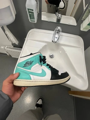 Turkosa och vita Air Jordan 1 High - Säljer ett par Air Jordan 1 High med turkosa, vita och svarta paneler. Skorna har klassisk Nike Swoosh på sidan, svart snörning och vit tåbox. Ovandelen är i läder och sulan är vit med svart yttersula. Riktigt snygga sneakers för dig som gillar streetwear.