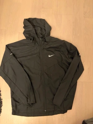 Nike miller jacka M/L - Svart vindjacka från Nike med dragkedja framtill och justerbar huva. Den är i L men den passar M. Jackan har en vit Nike-logga på bröstet och diskreta reflexdetaljer på ärmarna. Tillverkad i lätt och vindtätt material, perfekt för blåsiga dagar Skriv om ni har frågor eller funderingar!.