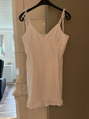 Vit kort klänning med spetsdetaljer - Söt vit kort klänning från Design By Si med smala axelband som knyts, v-ringning och broderade spetsdetaljer. Klänningen har en lätt volangkant nertill och dragkedja i ryggen. Perfekt för sommarens alla tillfällen.