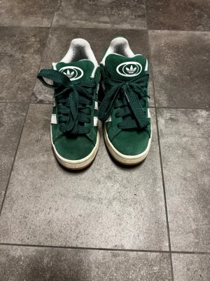 Gröna Adidas sneakers i mocka - Mörkgröna Adidas sneakers i mocka med vita detaljer och klassiska tre ränder på sidan. Skorna har gröna skosnören,gul  sula med vit rand och Adidas-logga på plösen. Lågt skaft och retrovibe som passar till streetwear.
