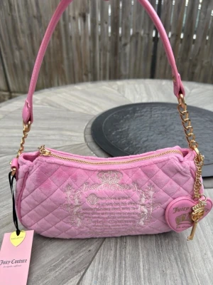 Rosa quiltad handväska Juicy Couture - Säljer en rosa quiltad handväska från Juicy Couture i mjuk velour med guldfärgade detaljer och kedja. Väskan har broderat emblem framtill, dragkedja med hjärtformad berlock och matchande axelrem. Perfekt för dig som gillar Y2K-stil och vill sticka ut.