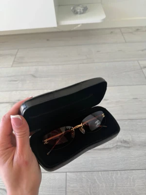 Chanel solglasögon med gulddetaljer - Snygga Chanel solglasögon med rektangulära, bruna tonade glas och guldfärgade detaljer. Bågarna är minimalistiska och har den klassiska Chanel-loggan på sidorna. Kommer med svart hårt fodral.