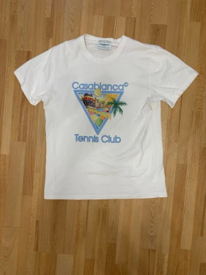 Vit Casablanca Tennis Club t-shirt - Snygg vit t-shirt från Casablanca med färgglatt tryck på bröstet som visar ett triangulärt motiv med palmer, tennisboll och texten 'Casablanca Tennis Club'. Klassisk passform och rund halsringning. Perfekt för dig som gillar sportig och trendig stil.