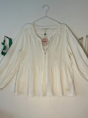 Vit långärmad blus från Odd Molly XXL - Säljer en vit långärmad blus från Odd Molly med smockade detaljer och diskret prickmönster. Blusen har rundad V halsringning med spetskant och knytband framtill. Materialet är lätt och luftigt, perfekt för en avslappnad look.