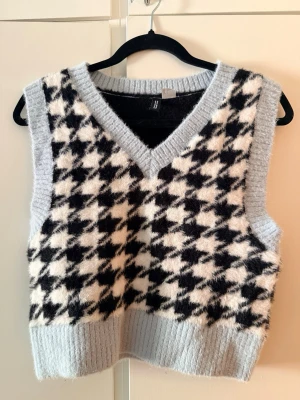 Rutig stickad väst från H&M Divided - Säljer en mysig stickad väst från H&M Divided i storlek S. Västen har ett rutigt mönster i svart och vitt med ljusblå ribbade kanter och v-ringning. Perfekt att styla över en skjorta eller t-shirt.