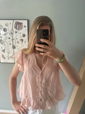 Ljusrosa volangblus med spetsdetaljer - Söt ljusrosa blus med volanger och spetsdetaljer längs ärmar och nederkant. Blusen har knappar framtill och en lös, luftig passform. Perfekt för en romantisk och feminin stil.