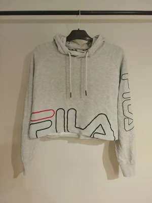 Grå cropped hoodie från FILA Nyskick - Snygg grå cropped hoodie från FILA med stor logga i svart och rött på magen och ärmen. Hoodien har huva med dragsko och långa ärmar. Perfekt för en sportig och avslappnad stil. Nyskick