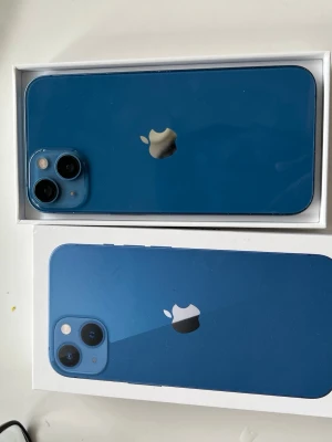 Blå Apple iPhone 13 med dubbla kameror - Snygg blå iPhone 13 med stilren design och dubbla kameror på baksidan. Telefonen har en platt baksida i glas och aluminiumram. Apple-loggan är centrerad och kamerorna sitter i ett hörn. Perfekt för dig som gillar modern teknik och cool färg. batteri 85%
