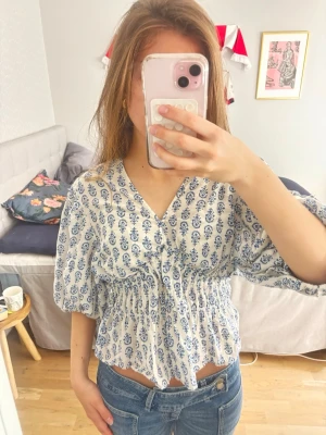 Vit och blå blus med v-ringning - Söt blus i vitt med blått blommigt mönster och v-ringad hals. Modellen har korta puffärmar, smockad midja och volangkant nedtill. Jag har storlek L men är storlek S i alla andra toppar så den passar. Jag har dock sytt lite i v ringen så att den passar mig men det kan man ta bort om man vill🥰