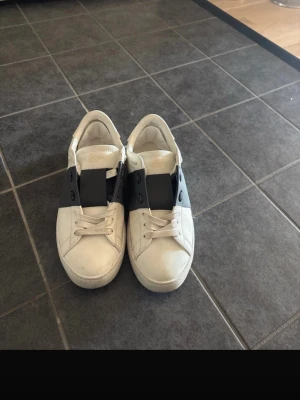 Vita sneakers med svart band Valentino - Snygga vita sneakers från Valentino med ett brett svart band över snörningen och nitar på hälen. Skorna har en klassisk låg profil och är tillverkade i läder. Perfekta för dig som vill ha en clean men ändå unik look.