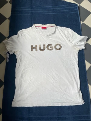 Vit HUGO t-shirt med rutigt tryck - Vit t-shirt från HUGO med korta ärmar och rund hals. Framsidan har ett stort rutigt HUGO-tryck i brunt och beige. Tillverkad i mjuk bomull för en skön känsla. Perfekt för en clean och trendig look.