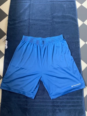 Blå träning shorts från Stanno - Snygga blå sportshorts från Stanno med elastisk midja och ledig passform. Perfekta för träning eller fotboll, tillverkade i ett lätt och snabbtorkande syntetmaterial. Diskret logga på ena benet för en clean look.
