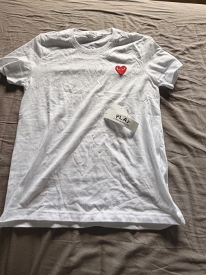 Vit Comme des Garçons PLAY t-shirt - Vit t-shirt från Comme des Garçons PLAY med det ikoniska röda hjärtat broderat på bröstet. Klassisk passform och rund halsringning. Tillverkad i mjuk bomull, perfekt för en clean och stilren look. Helt ny, prislapp är avtagen men följer med. Priset kan diskuteras.