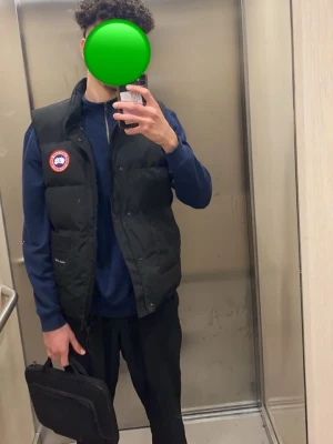 Svart dunväst från Canada Goose - Säljer en svart dunväst från med klassisk logotyp . Västen har hög krage, dragkedja och tryckknappar framtill samt två stora fickor. riktigt snygg 