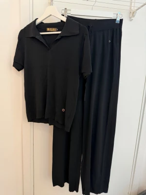 Svart loungewear-set från Loro Piana - Stilrent svart loungewear-set från Loro Piana med kortärmad topp med krage och vida byxor med resår i midjan. Mjukt och bekvämt material, perfekt för chill dagar hemma. Tillverkad i Italien.