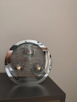 Rund glittrig ljushållare i glas - Säljer en rund ljushållare i glas och metall med glittrig bakgrund och plats för tre värmeljus. Modern design med spegelkant och små dekorstenar som ger extra bling. Perfekt som dekoration i vardagsrummet eller sovrummet.