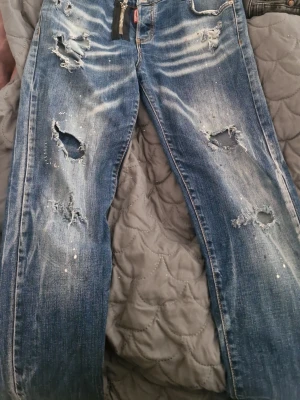 Blå slitna jeans med hål - Säljer ett par blå jeans med slitningar och stora hål på benen. Jeansen har en tvättad look med ljusa partier och små färgstänk. Klassisk femficksmodell och rak passform. Perfekta för en avslappnad och trendig stil.