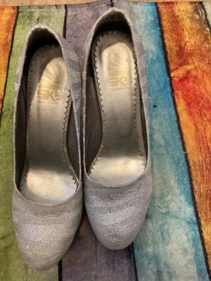 Silvriga pumps från SKL - Snygga silvriga pumps från SKL med glittrig finish och rund tå. Skorna har en klassisk form och är tillverkade i ett skimrande textilmaterial som ger en festlig känsla. Perfekta för dig som vill sticka ut med stilrena detaljer.