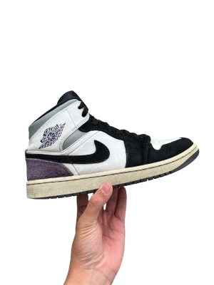 Nike Air Jordan 1 Mid svart/vit/lila - Nike Air Jordan 1 Mid sneakers i svart, vit och lila mocka och läder. Klassisk Swoosh-logga på sidan och Jordan Wings-märke vid ankeln. Grå detaljer runt kragen och vintage-inspirerad gulnad sula. Perfekt för dig som gillar streetwear och snygga detaljer.