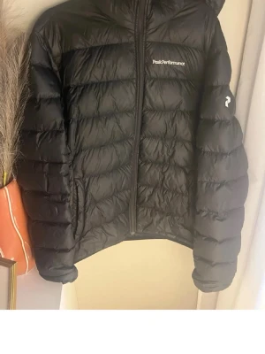 Svart dunjacka Peak Performance - Snygg svart dunjacka från Peak Performance med huva och dragkedja framtill. Jackan har diskret logga på bröstet och ärmen, samt quiltad design som ger en sportig look. Perfekt för kalla vinterdagar och riktigt varm tack vare dunfyllningen. ⚠️dragkedjan är inte trasiga den är svår att stänga så jag klassar den som varning ⚠️ 
