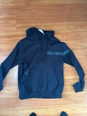 Hugo boss hoodie - Säljer en svart hoodie med dragkedja och huva. Den har två stora fickor framtill och en diskret grå reflexdetalj över bröstet. Hoodien har ribbade muddar och snören i huvan. Perfekt för en avslappnad och cool look.