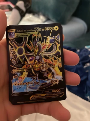 Pokémonkort VMAX God Of Pokémon - Unikt Pokémonkort med VMAX God Of Pokémon-design. Kortet har en svart och gul färgskala med holografiska detaljer och visar en kraftfull Pokémon i dynamisk pose. Texten på kortet är på engelska och det har en fantasifull, exklusiv layout.