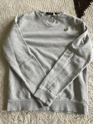 Grå sweatshirt från Lyle & Scott - Snygg grå sweatshirt från Lyle & Scott med klassisk rund halsringning och det ikoniska gula logomärket broderat på bröstet. Tröjan har långa ärmar och ribbade muddar vid ärmslut och nederkant. Perfekt för en avslappnad och stilren look.