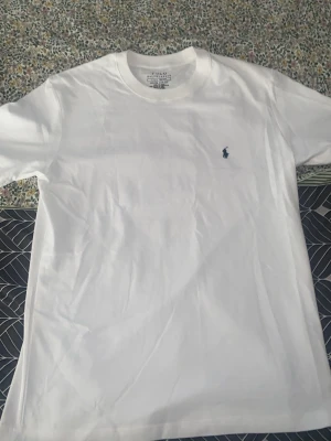 Vit slim fit t-shirt från Polo Ralph Lauren - Klassisk vit t-shirt från Polo Ralph Lauren i 100% bomull. Slim fit-modell med korta ärmar och rund halsringning. Diskret blå logga broderad på bröstet ger en stilren look. Perfekt basplagg för alla tillfällen.