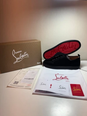 Svarta sneakers från Christian Louboutin - Snygga svarta sneakers från Christian Louboutin med klassisk röd sula och diskret logga på sidan. Skorna har en låg profil, svarta skosnören och ovandel i mocka. Perfekta för dig som vill ha en exklusiv och stilren look.