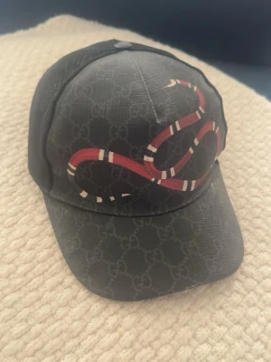 Gucci  keps  - Snygg svart keps från Gucci med klassiskt GG-mönster och en röd-vit orm på framsidan. Kepsen har mesh på sidorna och justerbar rem bak. Materialet är en mix av canvas och mesh vilket ger en cool och luftig känsla. Lite skador här och där men annars mycket bra skick och så klart äkta ✅